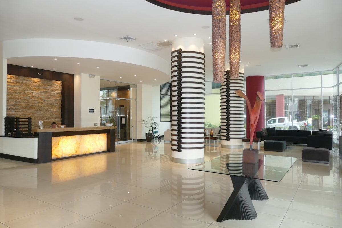 The lobby area features a modern design with polished tile flooring and soft lighting. A reception desk is positioned on the left, complemented by decorative elements such as pendant lighting and elegant furnishings. Large windows bring natural light into the space, enhancing the inviting atmosphere.