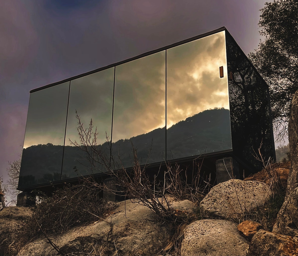 A modern structure is set against a rocky landscape, featuring reflective glass panels that mirror the surrounding mountain scenery and cloudy sky. The elevated design sits on supports, surrounded by natural vegetation and boulders, emphasizing the connection with the landscape.