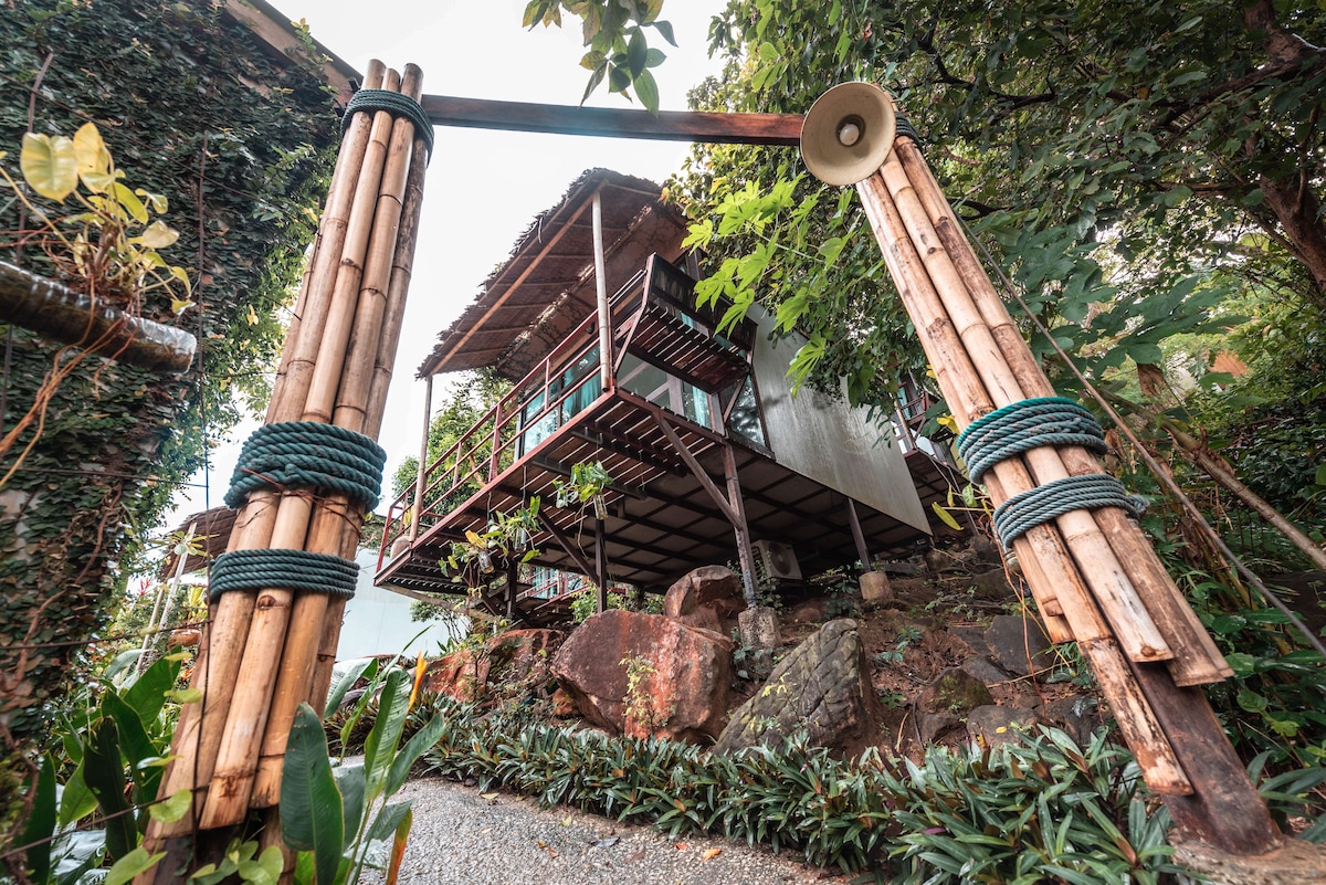 A rustic bungalow is perched on a hillside, framed by lush greenery and tropical plants. The structure is supported by a sturdy bamboo entrance, while a wooden deck provides a vantage point over the surroundings. Natural stone accents complement the vibrant landscape.