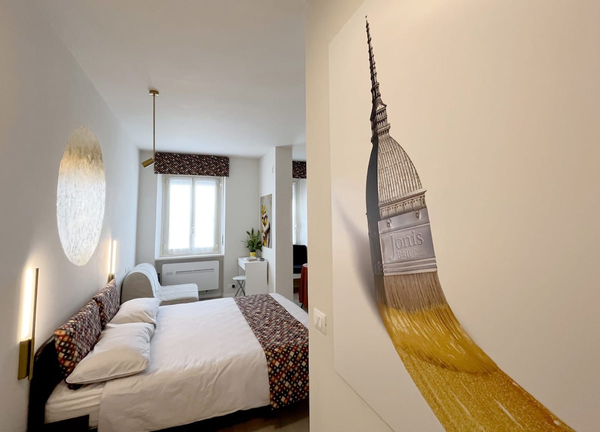 A spacious bedroom features a queen-size bed dressed in neutral linens. The wall behind holds a striking mural of a tower, complementing the room's modern decor. Natural light flows in through a window with patterned curtains, contributing to a bright and welcoming environment.