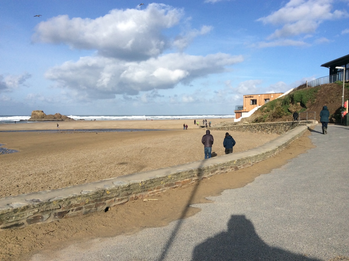 The image captures a sandy beach with gentle waves lapping at the shore. People are seen walking along the beach and path, with a beachfront building visible in the background. The sky features scattered clouds, adding a sense of openness to the coastal view.