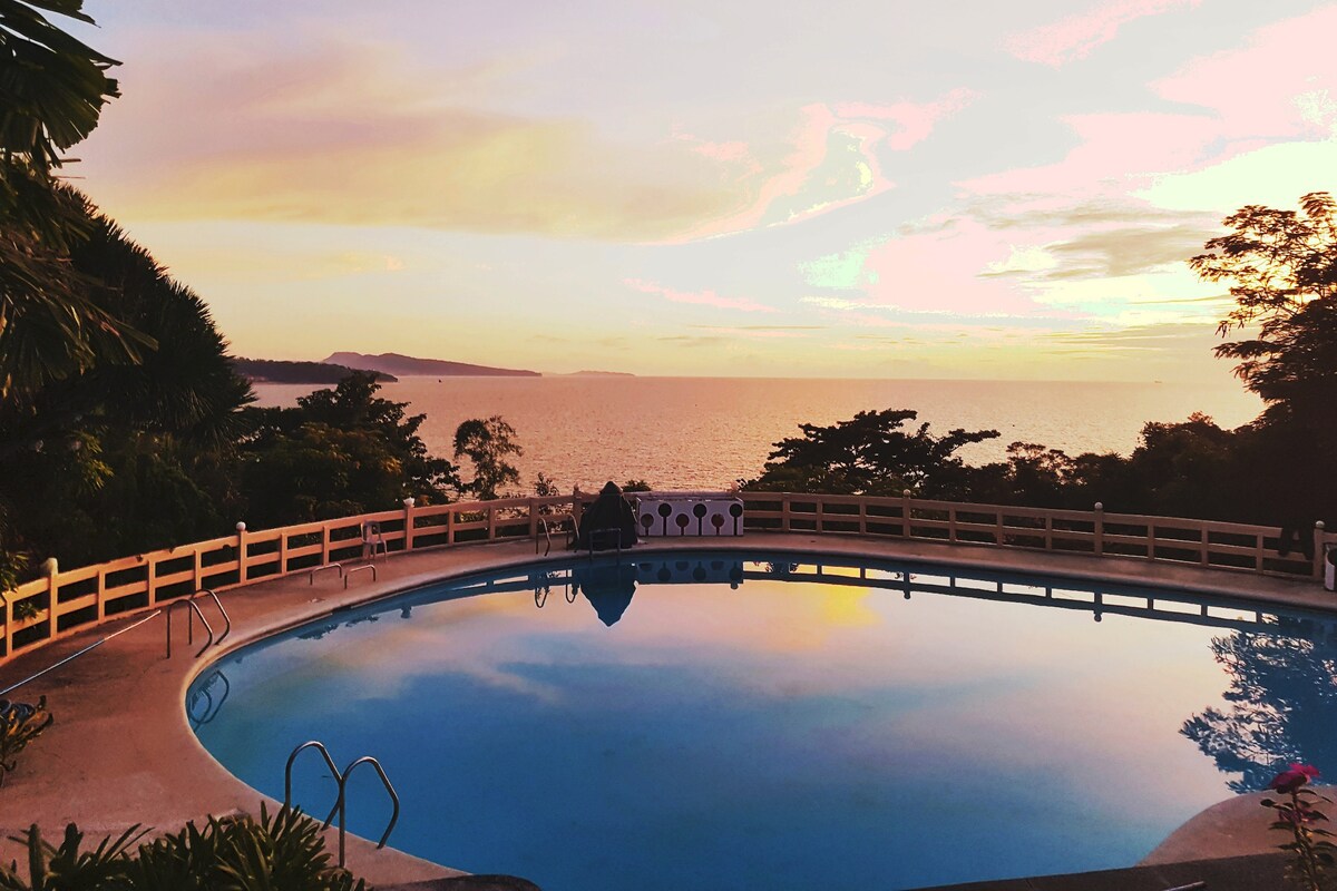 A round pool is positioned overlooking the ocean, reflecting hues of pink and orange from the sunset. Lush greenery surrounds the area, and a wooden railing enhances the view. The tranquil scene creates an inviting atmosphere for relaxation.