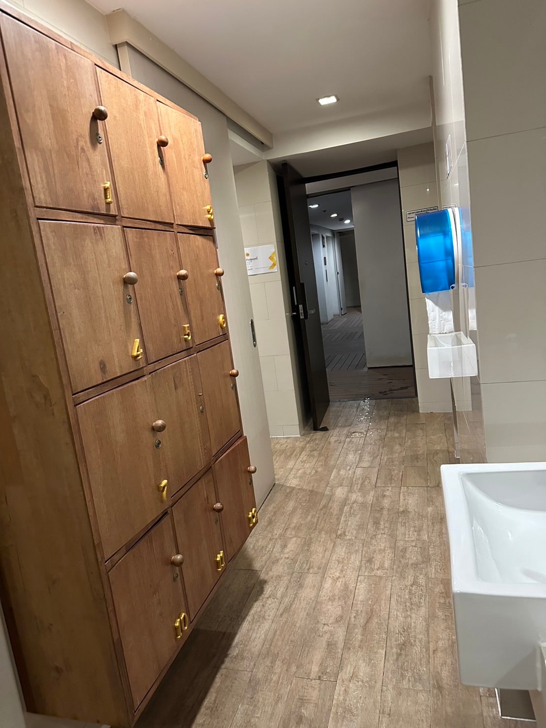 A clean and well-lit hallway features a wooden locker unit with numbered compartments along the left side. A white sink is positioned nearby, while a dark door leads to additional areas in the condo. Neutral tile and wood flooring create a warm, cohesive look.
