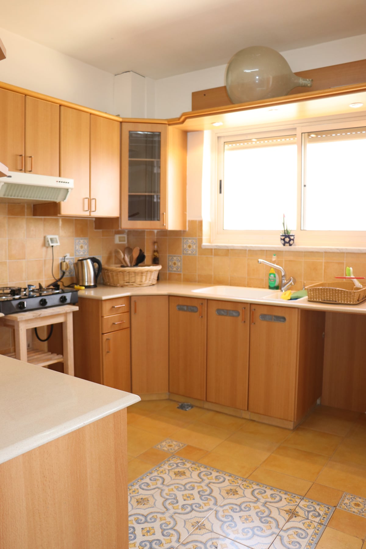 The kitchen features warm wood cabinetry complemented by tiled surfaces. A gas stove and various appliances, including a kettle, are visible. Natural light enters through a window, illuminating a functional layout with ample counter space and storage. A subtle decorative jar is placed on a shelf.