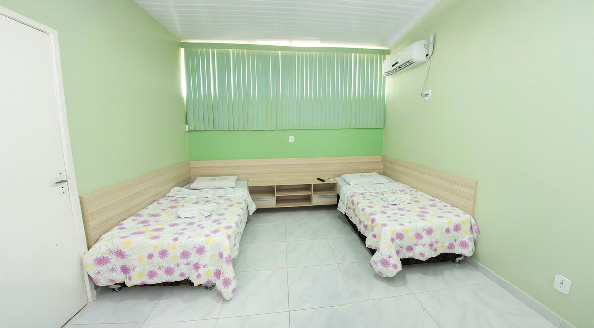 The room features two neatly arranged beds, each covered with floral-patterned linens. Light green walls create a calming atmosphere, while large windows with vertical blinds allow natural light to illuminate the space. A wall-mounted air conditioning unit is visible.