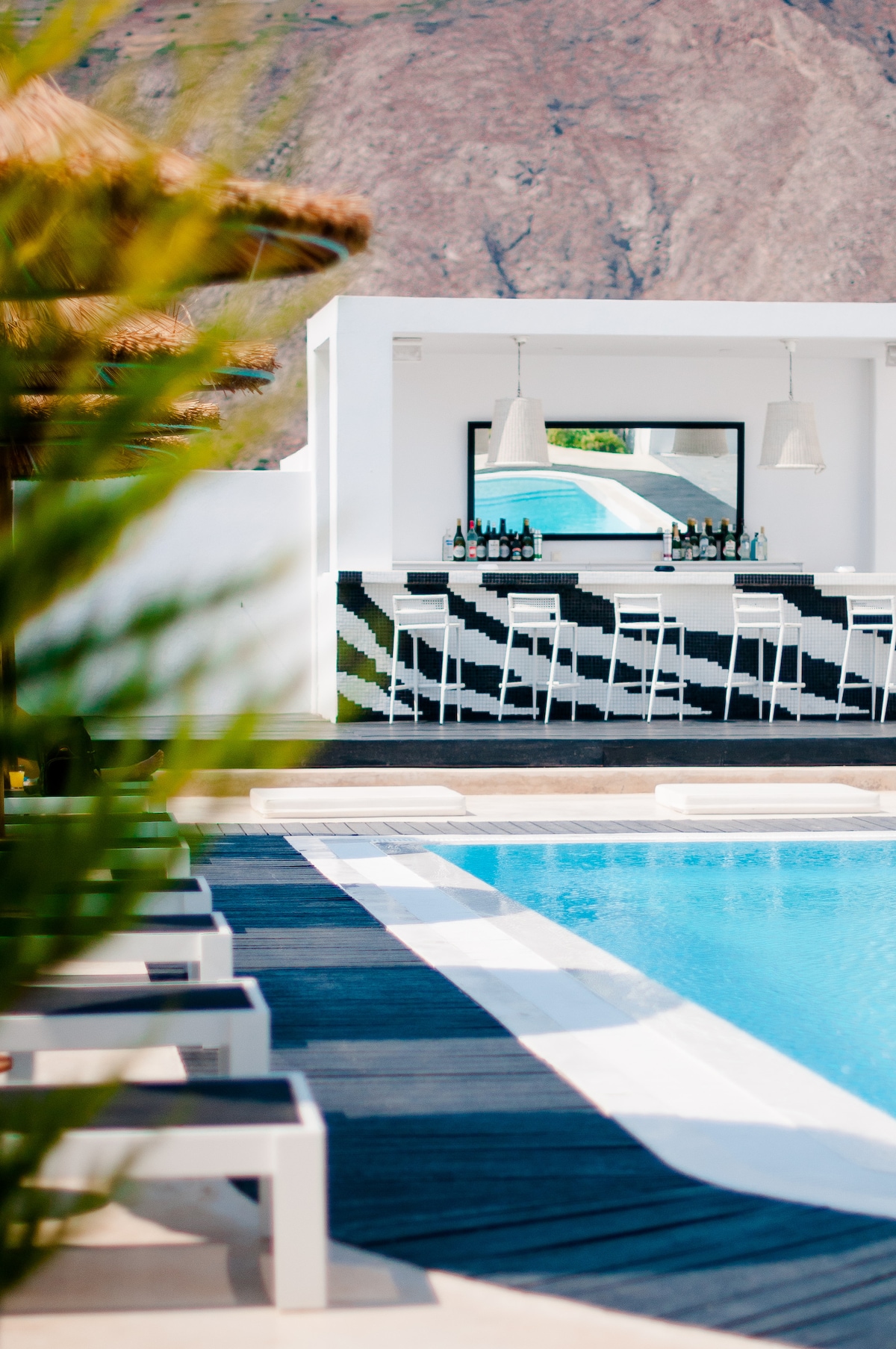 A modern outdoor bar area is situated beside a clear blue pool, featuring a stylish black and white striped counter. Two large pendant lights hang above the bar, and bar stools are arranged along the counter, offering a relaxed gathering space.