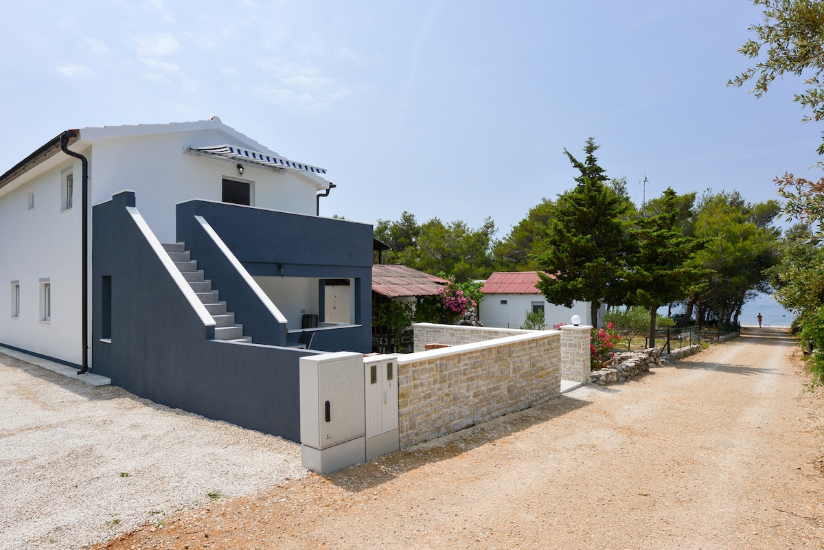 A modern white house with a blue accent stands at the end of a gravel path lined with trees and greenery. A flight of stairs leads to the upper level, while the beach is visible in the distance, framed by lush surroundings.