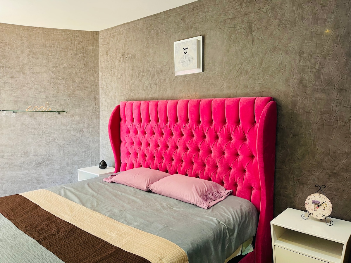A spacious bedroom features a striking pink tufted headboard above a neatly made bed. Two soft pillows rest against the headboard, and a light grey blanket covers the bed. Nightstands are positioned on either side, contributing to a cozy yet modern decor.