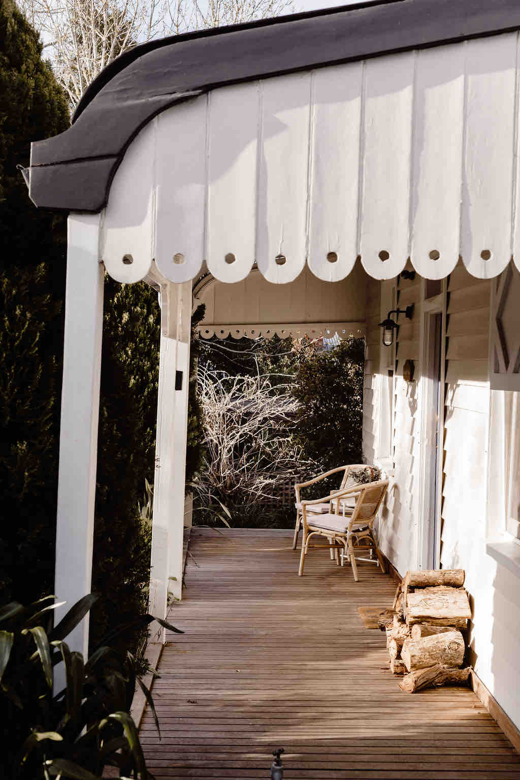 The entrance features a sheltered verandah with a white awning. A wooden bench is positioned beside a stack of firewood, offering a welcoming space to relax. Surrounding greenery enhances the sense of privacy and tranquility.