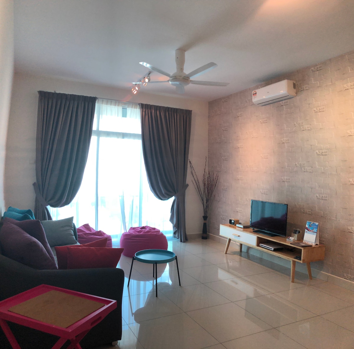 A spacious living area features large windows with sheer curtains, providing ample natural light. Comfortable seating arrangements include a sectional sofa with colorful pillows and two bean bag chairs. A flat-screen TV is positioned on a wooden console, while a small table adds a decorative touch.