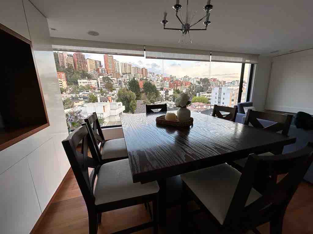 A spacious dining area is highlighted by a large wooden table surrounded by six upholstered chairs. A decorative centerpiece adorns the table. Expansive windows offer a view of the city skyline, allowing natural light to fill the space.