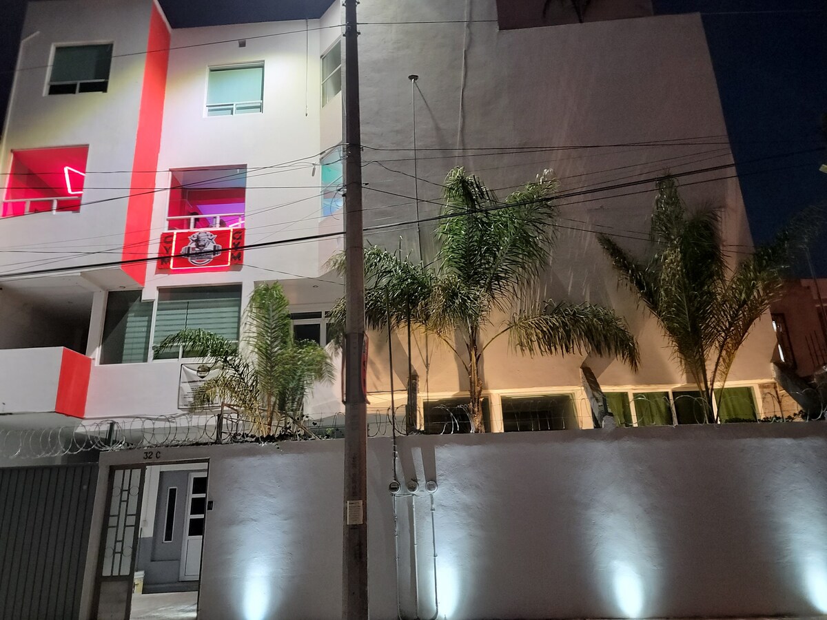 The three-story building is positioned under night lighting, showcasing a modern facade with vibrant red accents. Palm trees are visible flanking the entrance, and soft illumination highlights the exterior walls, creating a welcoming ambiance.