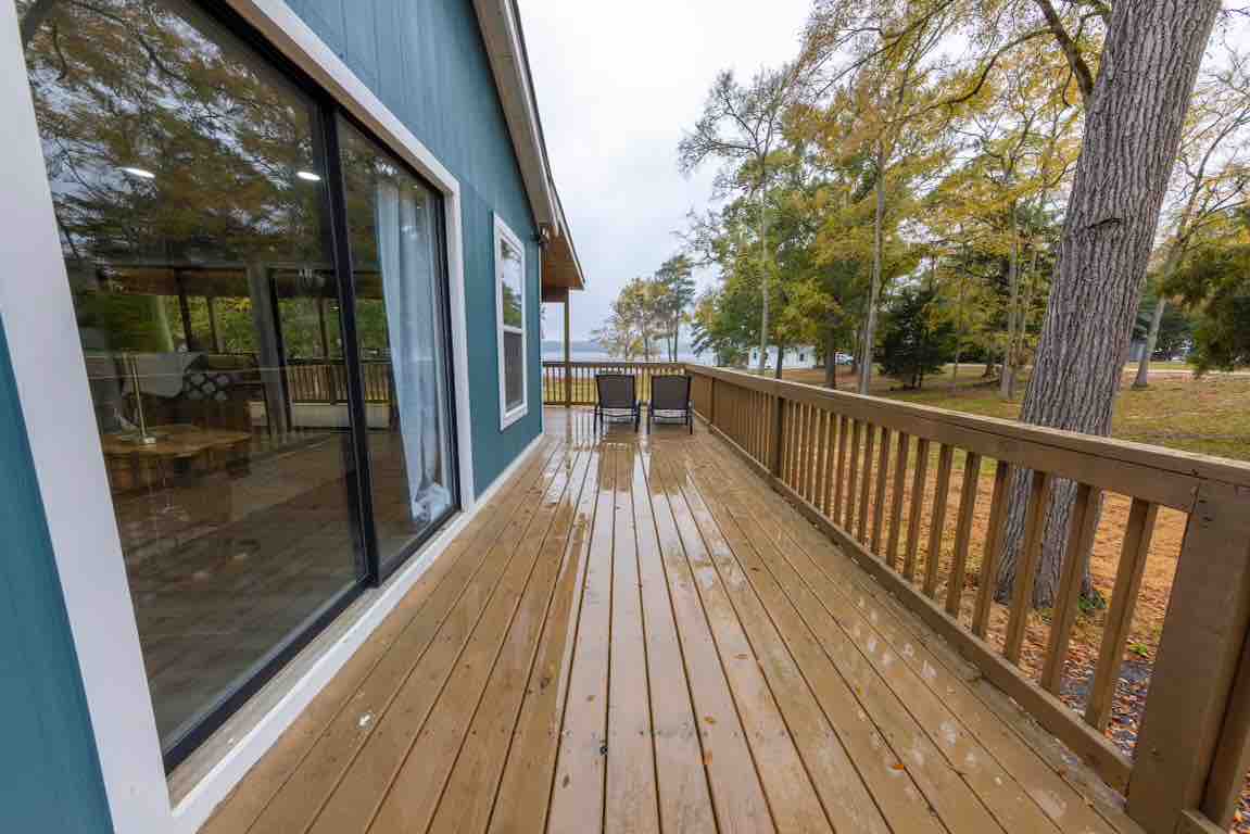 A spacious deck extends from the cottage, lined with wooden railing. It is partially wet, reflecting the surroundings. Trees with autumn foliage are visible in the background, along with expansive views of Lake Wateree, providing a serene outdoor space.
