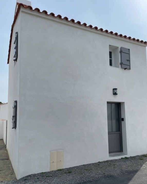 A modern, two-story white house with a simple exterior design and a terracotta-tiled roof is depicted. The front door is framed by a sleek gray entrance, complimented by evenly spaced window shutters, adding to the clean aesthetic of the property.