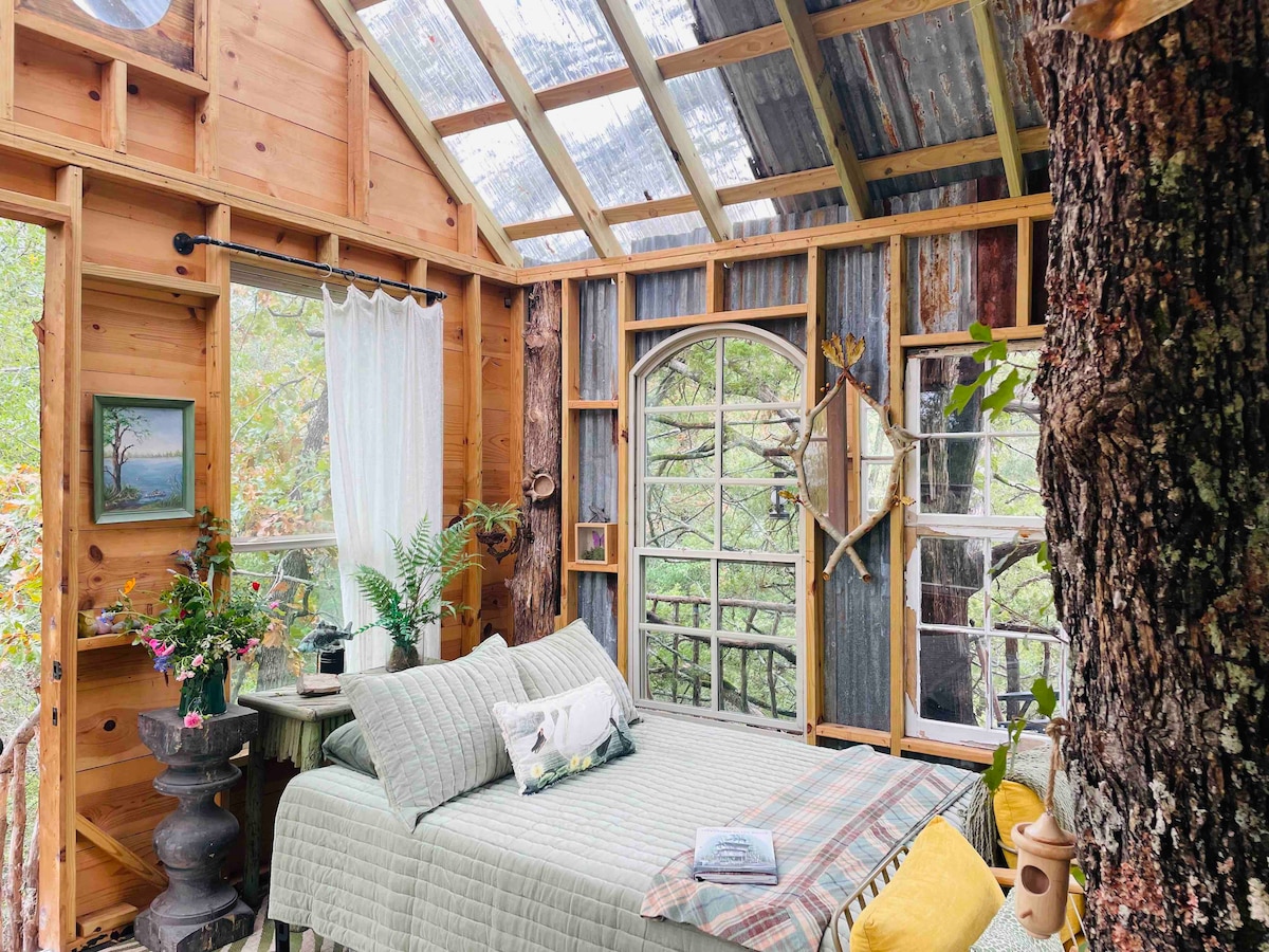 A cozy sleeping area surrounded by natural wood features large windows offering views of the outdoors. A spacious bed is adorned with a patterned blanket, and decorative plants and artwork enhance the rustic appeal of the space.