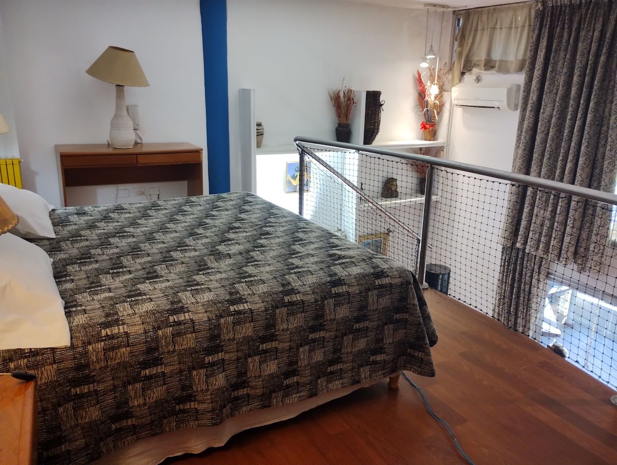 A loft-style bedroom features a spacious bed with a patterned blanket. A wooden bedside table holds a lamp. Soft lighting complements the warm tones of the wood flooring. Decorative elements are visible in the background, enhancing the serene ambiance.