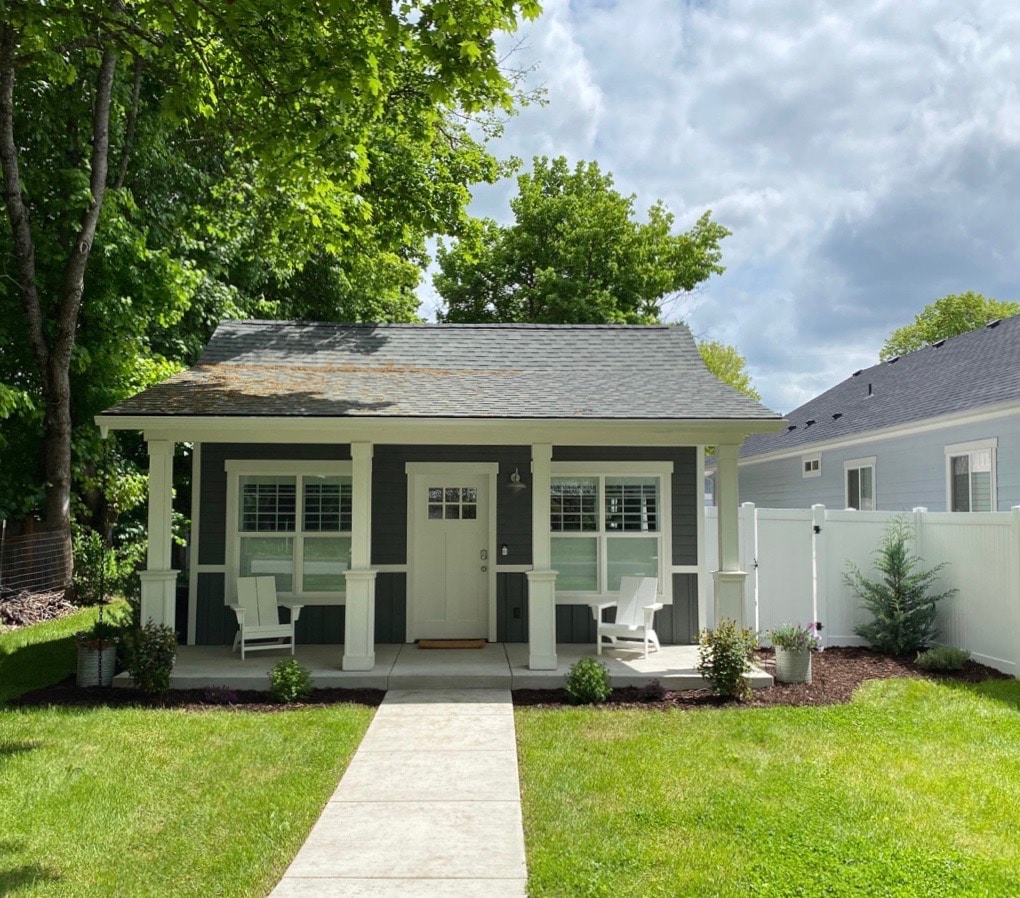 A charming bungalow is featured, showcasing a light grey exterior with white trim. The front porch is adorned with comfortable seating, framed by lush green trees. A concrete pathway leads to the welcoming entrance, enhancing the inviting appearance of the home.