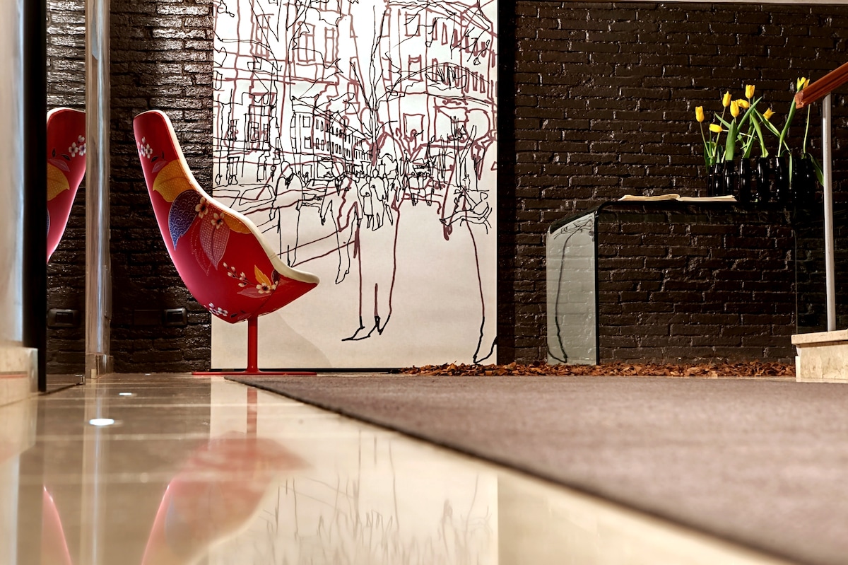 A warm and inviting lobby area is displayed, featuring a colorful chair alongside a minimalist glass table. A patterned wall mural depicting a city scene complements the modern decor, while fresh flowers in a long vase add a touch of nature. The polished floor reflects the surroundings.