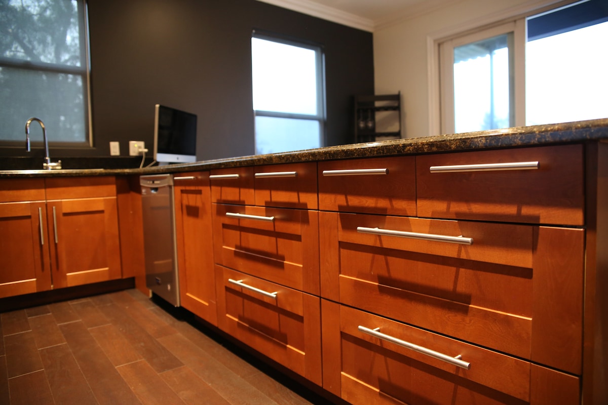 The kitchen features modern wooden cabinetry with sleek silver handles and a granite countertop. A large island is centrally positioned, enhancing workspace. A small window allows natural light, while dark walls provide contrast. The space is designed for both functionality and comfort.