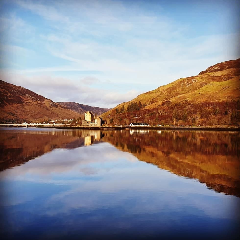 A calm loch reflects the surrounding hills and a historic castle in the distance. The landscape features gentle slopes adorned with various shades of brown and green, while the sky displays soft blue tones with scattered clouds.