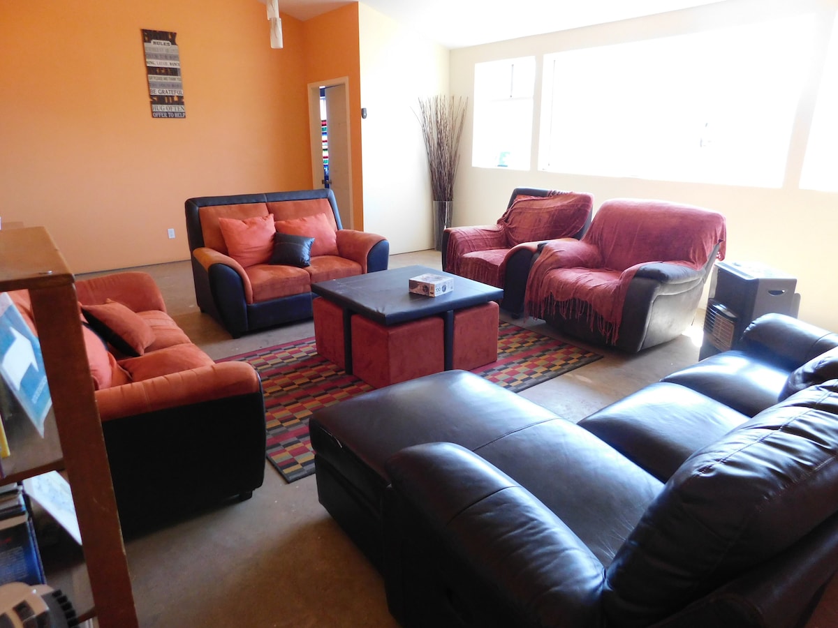 A communal lounge area is furnished with multiple seating options, including two armchairs and three sofas in varying shades of red and black. A central coffee table is positioned on a patterned rug, with large windows allowing natural light to fill the space.