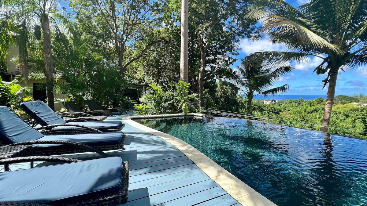 A sunlit infinity pool with a tranquil blue surface extends into the landscape, surrounded by lush greenery and tropical palm trees. Lounge chairs with blue cushions are positioned along the pool edge, offering a relaxing spot for guests to enjoy the serene view of the ocean.