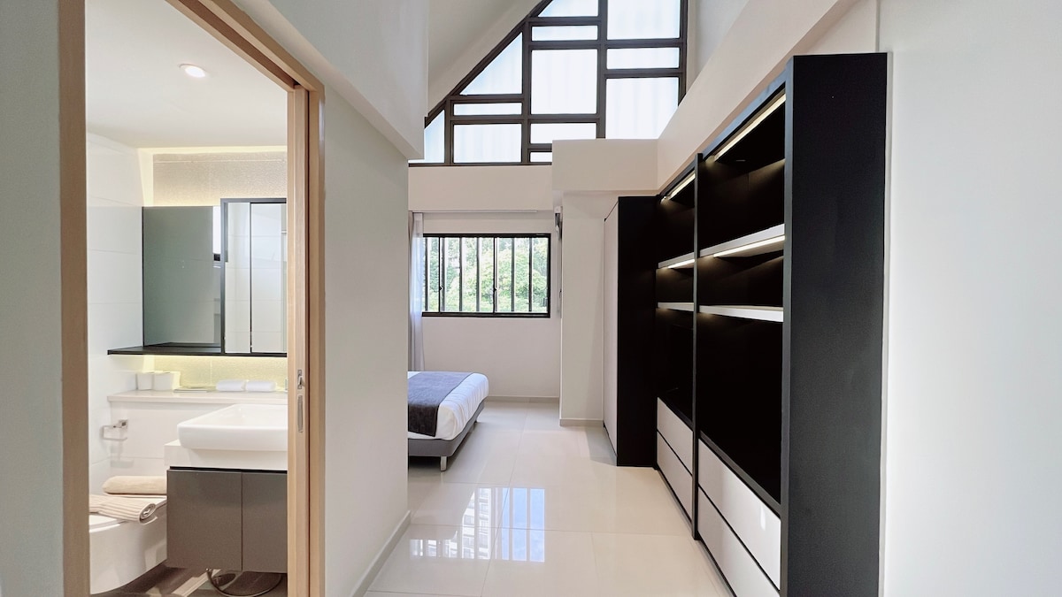 The bright and spacious room features high ceilings and large windows, allowing natural light to fill the space. A comfortable bed is positioned against one wall, while sleek shelving units offer storage. A modern private bathroom is visible through an open doorway.