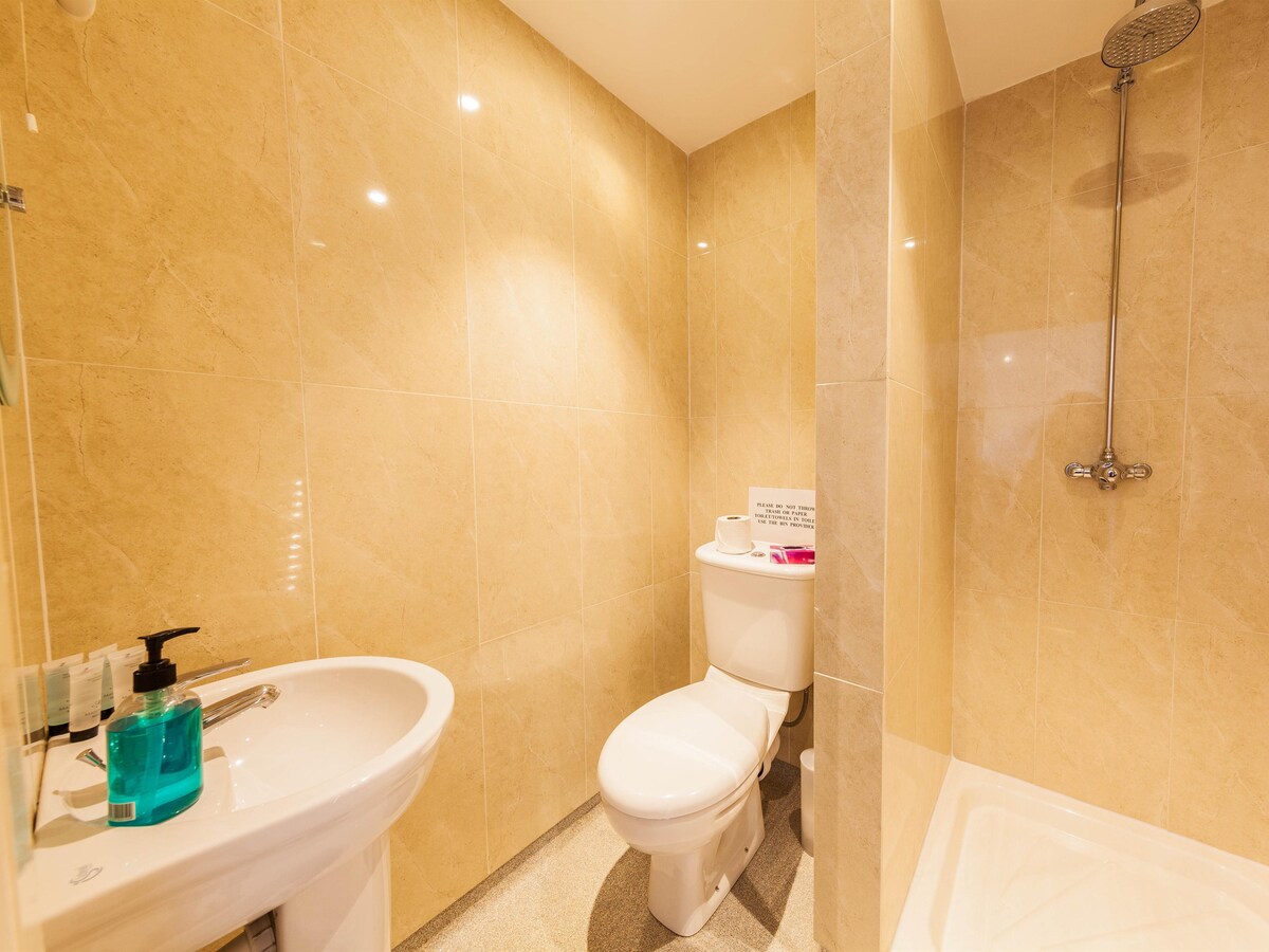 A modern bathroom features light-colored tiles on the walls and floor. A sink is positioned next to a toilet, with toiletries and a plastic bottle placed nearby. A shower area is present, equipped with a wall-mounted shower head.