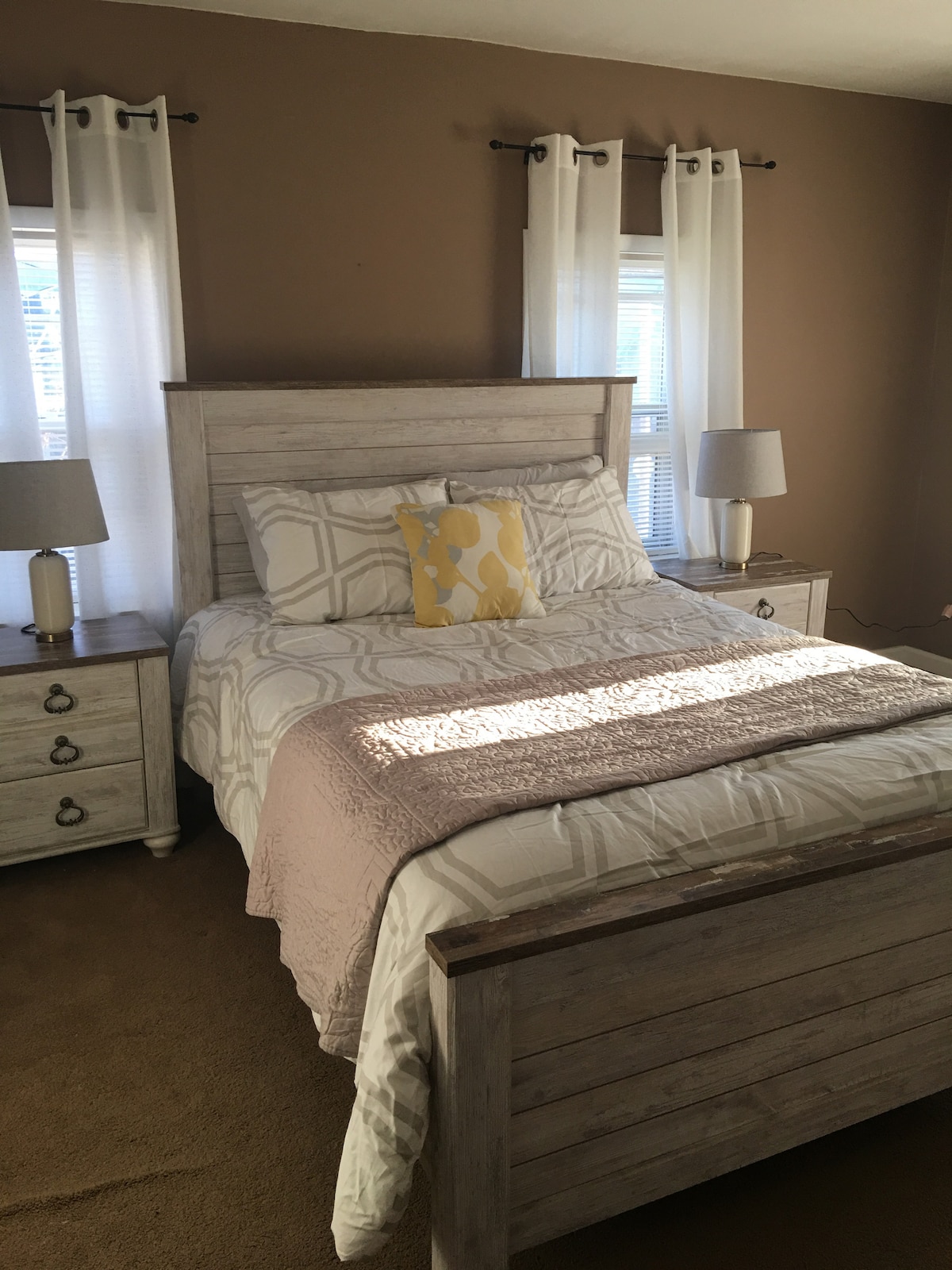 A bright bedroom features a spacious bed framed in light wood, adorned with a neutral patterned quilt and a decorative yellow pillow. Two bedside tables, each with a lamp, flank the bed, while natural light filters through sheer white curtains over dual windows.