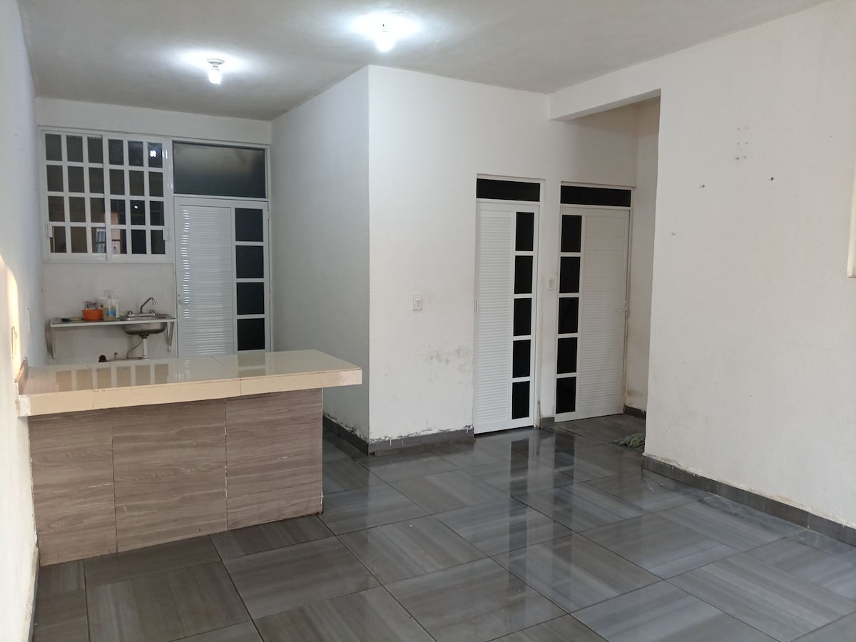 An open living area features a sleek tile floor and neutral walls. A light-colored countertop separates the kitchen area from the space, while multiple doors to the left lead to separate rooms. Natural light fills the area through multiple windows.