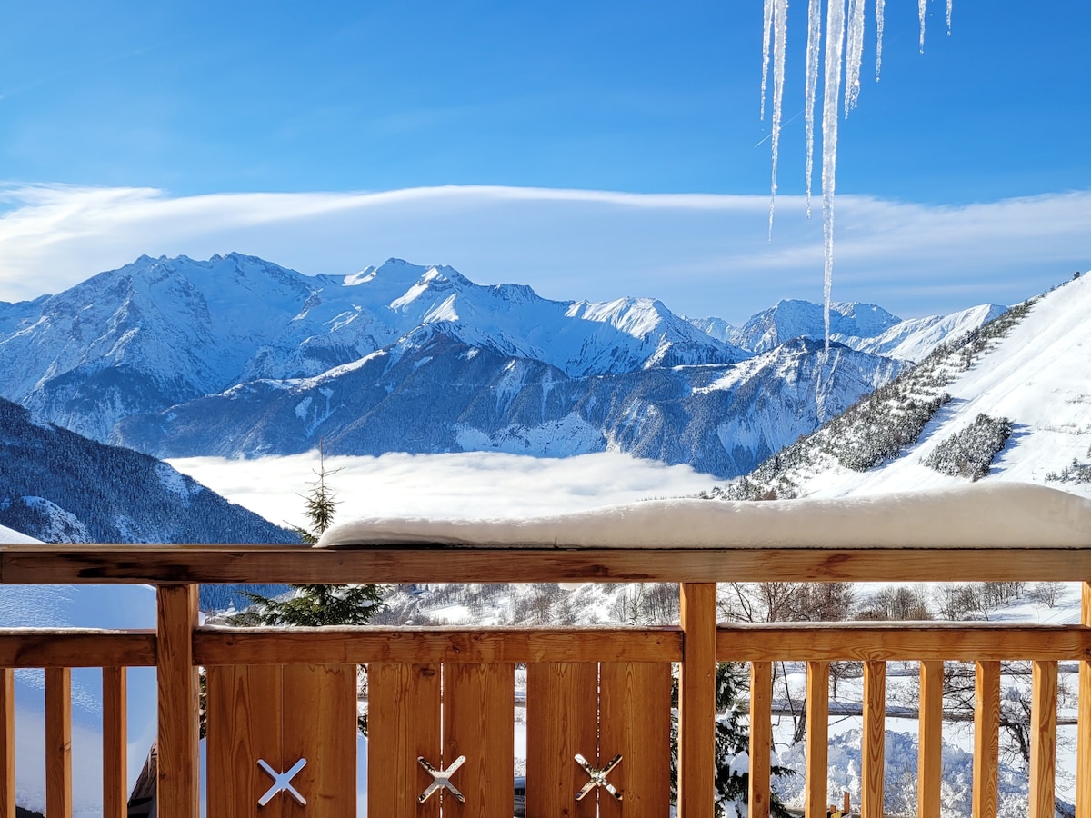 A balcony railing constructed of wood overlooks a breathtaking mountain landscape. Snow-capped peaks rise majestically in the background under a clear blue sky. Icicles hang from the edge of the balcony, adding a winter touch to the serene view.