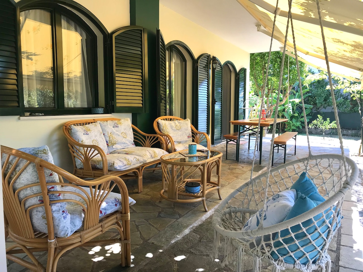 A shaded outdoor seating area features a comfortable rattan sofa set with floral cushions and a coffee table. A hanging swing chair adds a playful touch, while a dining table is set nearby, surrounded by lush greenery.