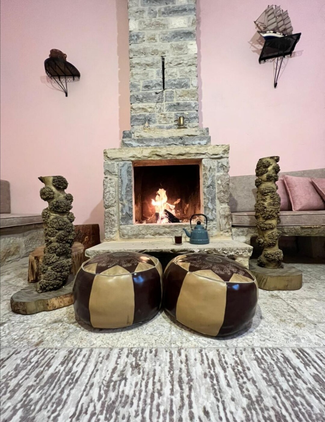 A warm living space features a rustic stone fireplace with a flickering fire. Two ornamental poufs, crafted with a unique design, are placed in front of the fireplace. The walls are painted in soft pink, complementing the natural stone elements.