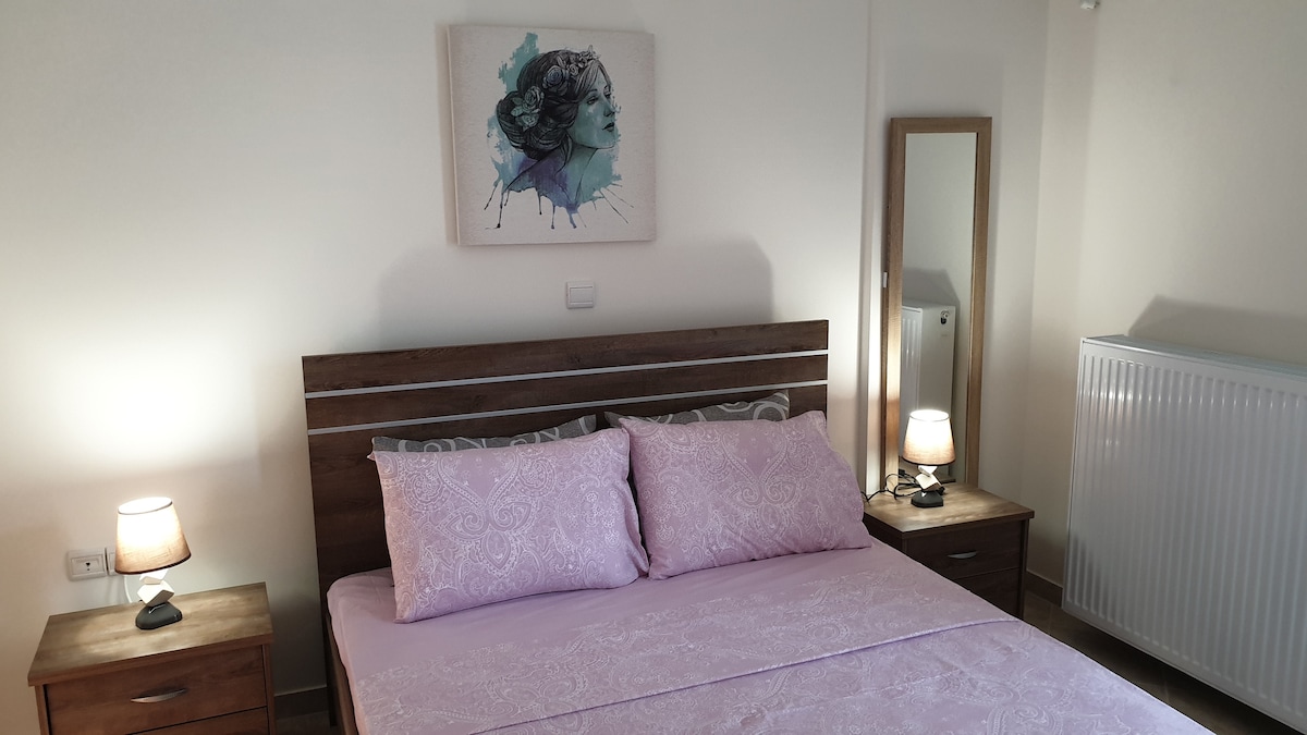 A well-lit bedroom is featured, showcasing a spacious bed adorned with soft pink bedding and matching pillows. Two bedside tables, each with lamps, are situated on either side. A large mirror reflects the room's light, and a piece of abstract wall art adds a pop of color.