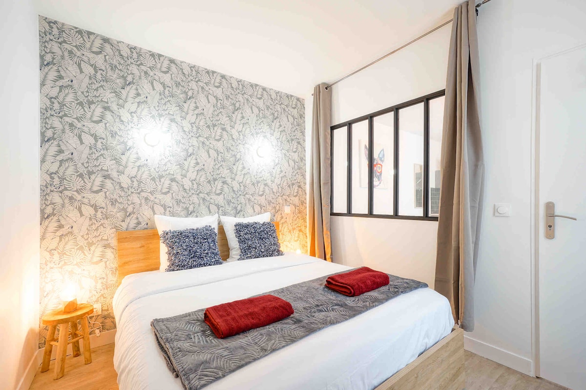 A well-lit sleeping area features a queen size bed dressed with crisp white linens and decorative pillows. Soft lighting from wall sconces enhances the relaxing atmosphere, while a patterned accent wall and large window allow for natural light to flow in.