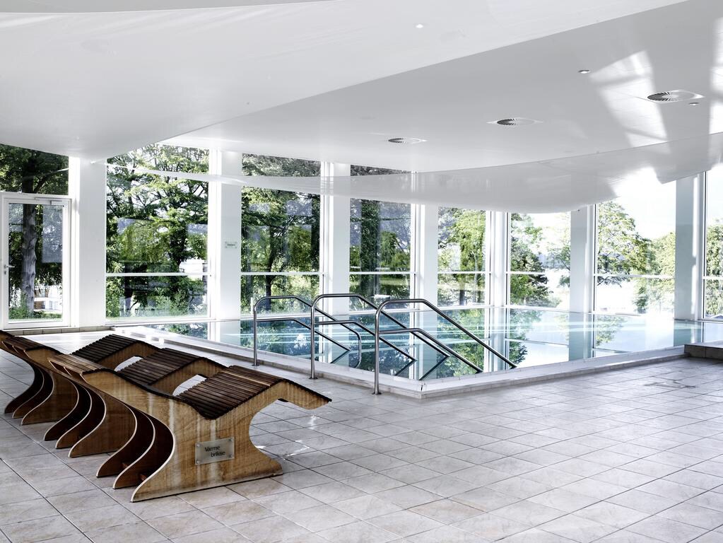 A bright indoor pool area is highlighted by large windows, allowing natural light to fill the space. Wooden lounge chairs are arranged neatly nearby, providing a comfortable place to relax. The pool features a gentle slope entry with safe railings.