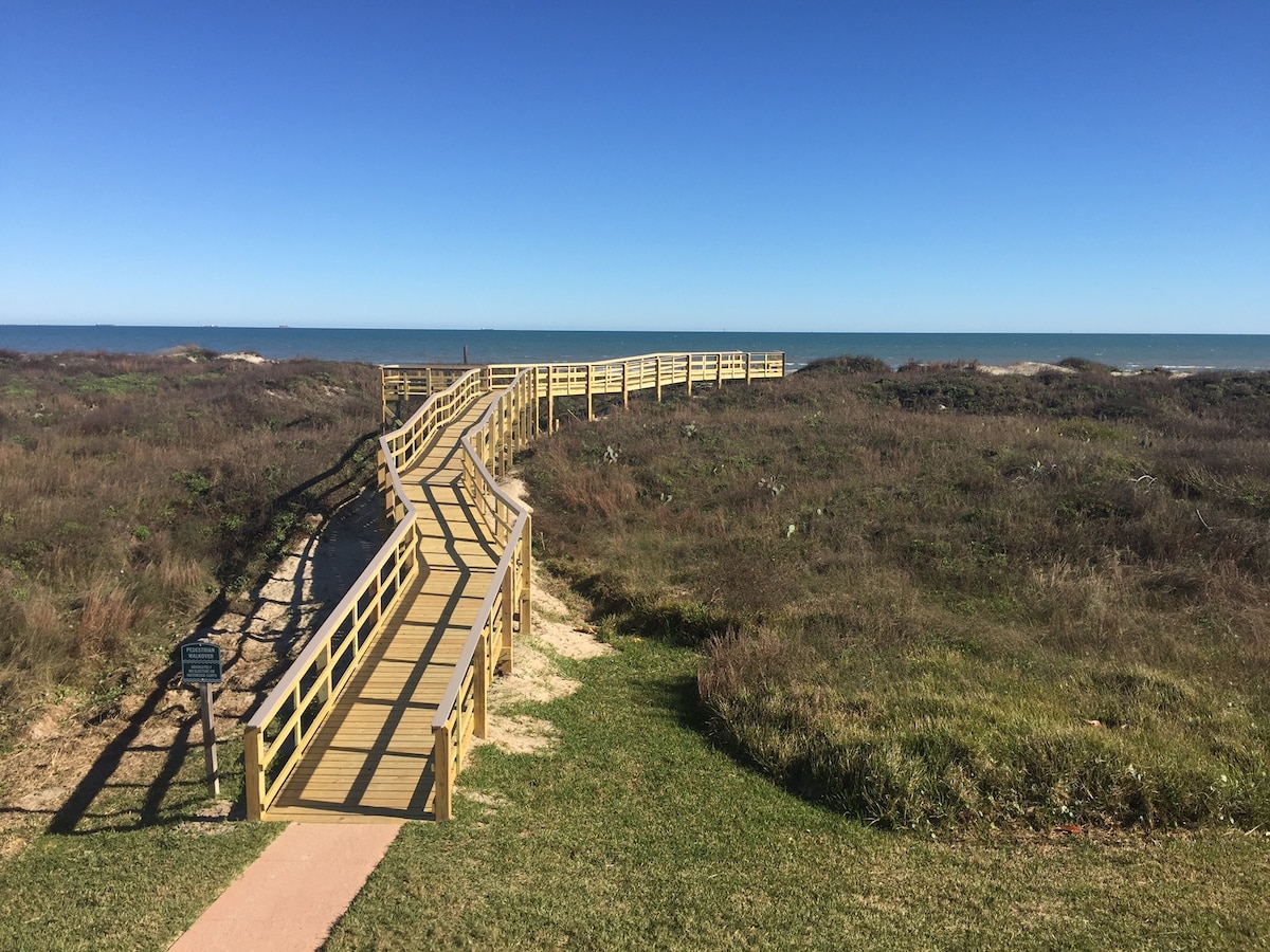 A winding wooden boardwalk extends across the landscape, leading to the beach and ocean. Lush green grass and brown coastal vegetation frame either side, while the clear blue sky meets the water in the distance, creating a serene and inviting pathway.