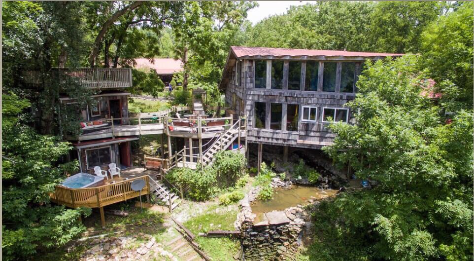 An aerial view of the property reveals a spacious deck surrounding a hot tub, nestled among lush greenery. The multi-level building showcases large windows, inviting natural light. A creek flows beside the structure, enhancing the natural setting.