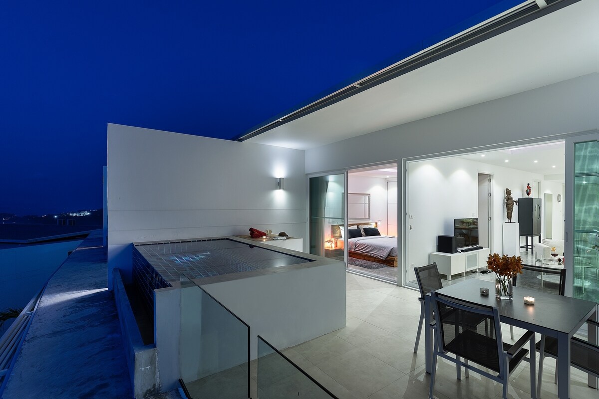 An expansive terrace showcases a private plunge pool with a serene water feature. A dining table with modern chairs is positioned on the left. Clear glass doors lead into the interior space, where soft lighting highlights an open living area that seamlessly integrates with the outdoor setting.