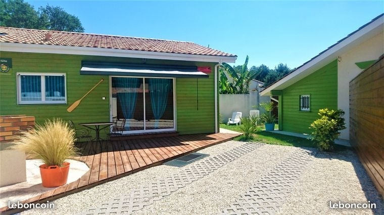 The exterior of a bright green gite is presented, featuring wooden decking and a covered terrace. A small table and chairs are visible on the patio, complemented by potted plants. A side view shows a landscaped area with a path leading to another structure.