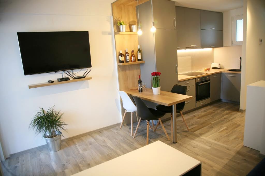 An open living space features a wall-mounted television above a minimalistic shelf. A dining table with black and white chairs is positioned next to the kitchen, which contains modern appliances. A vase of fresh flowers adds a touch of color, with warm lighting illuminating the area.