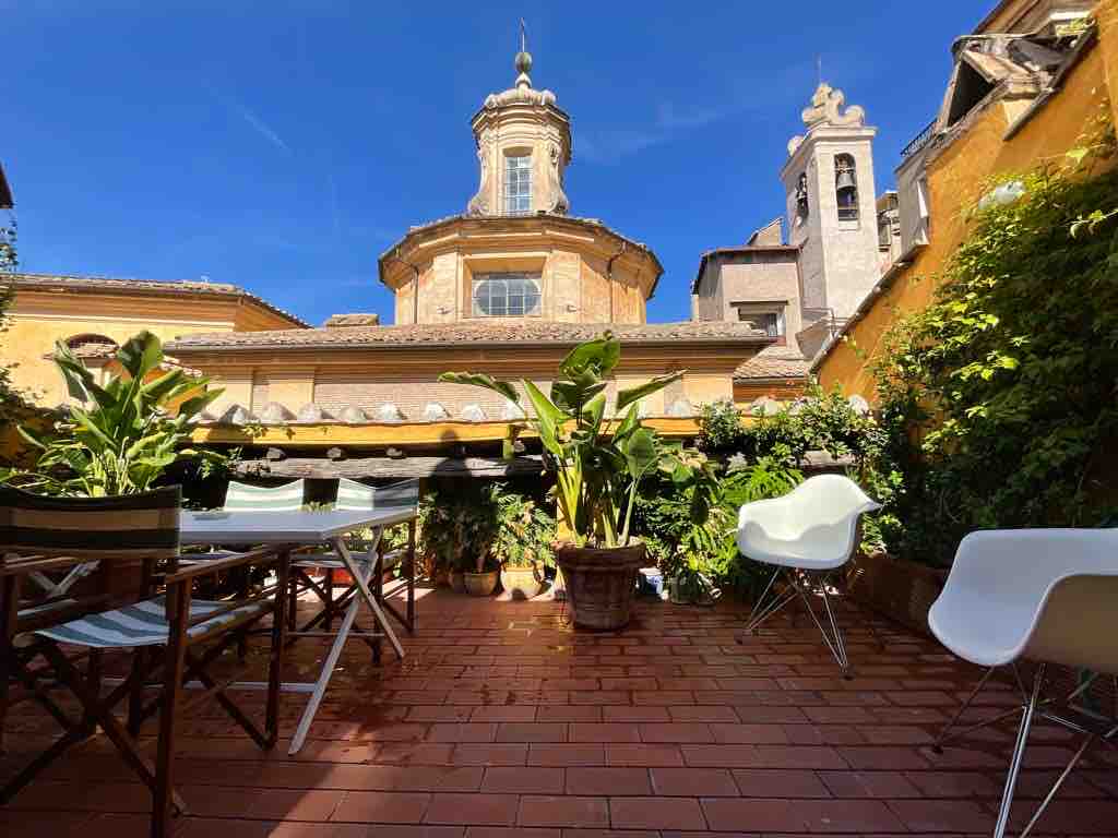 A private terrace is presented, featuring tiled flooring adorned with potted plants. A dining table with chairs is positioned along one side, offering a relaxing space. The backdrop includes the dome of a church, framed by vibrant greenery and warm yellow building facades.