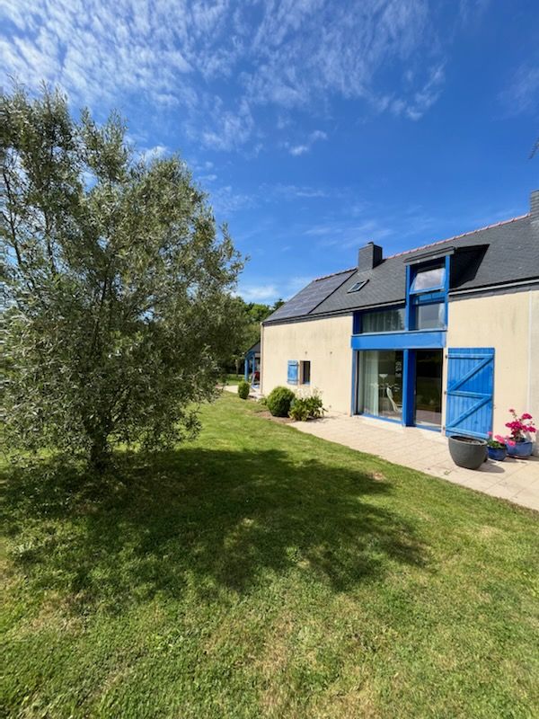 The exterior of a light-colored house is shown, complemented by blue window shutters. A grassy area surrounds the property, featuring a small tree and potted flowers. Clear skies and abundant sunlight create a bright and open atmosphere.