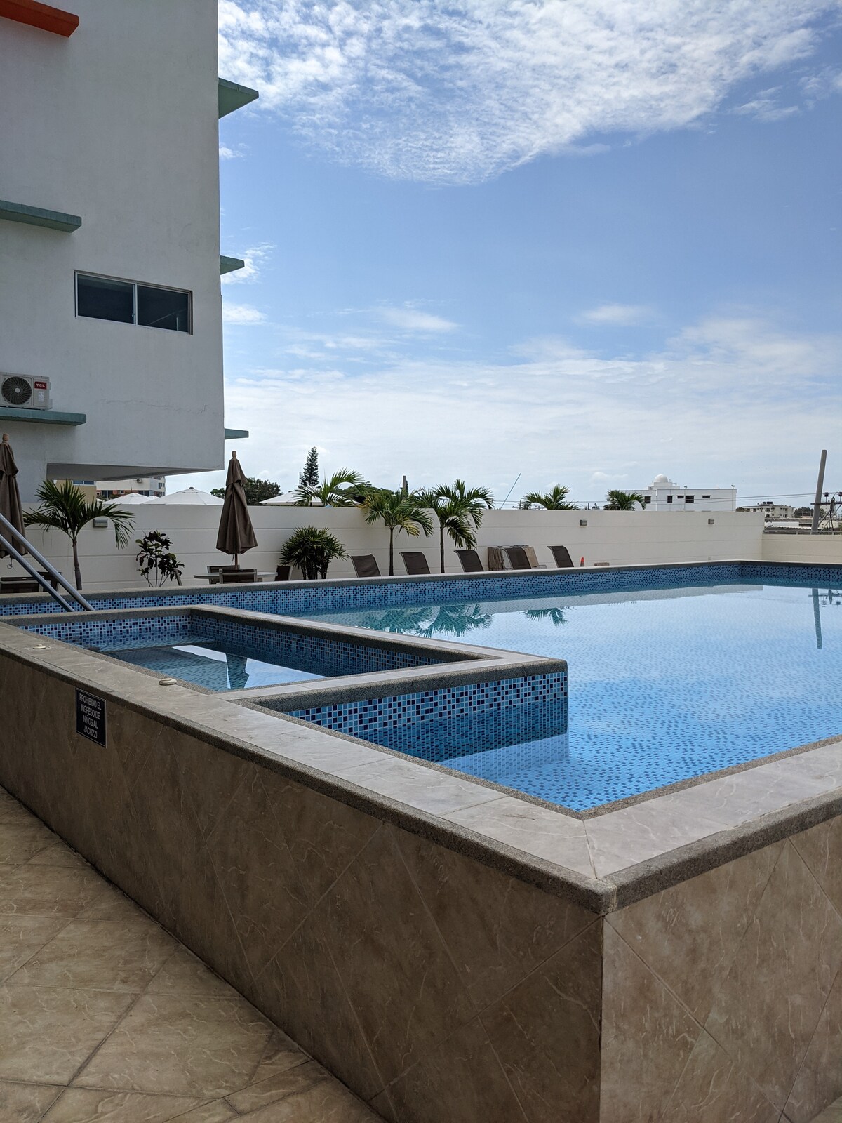 A clear view of a communal swimming pool featuring a shallow jacuzzi adjacent to it. Sun loungers with umbrellas are positioned along the poolside, while the surrounding foliage adds a touch of greenery. The sky is partly cloudy, providing a serene atmosphere.