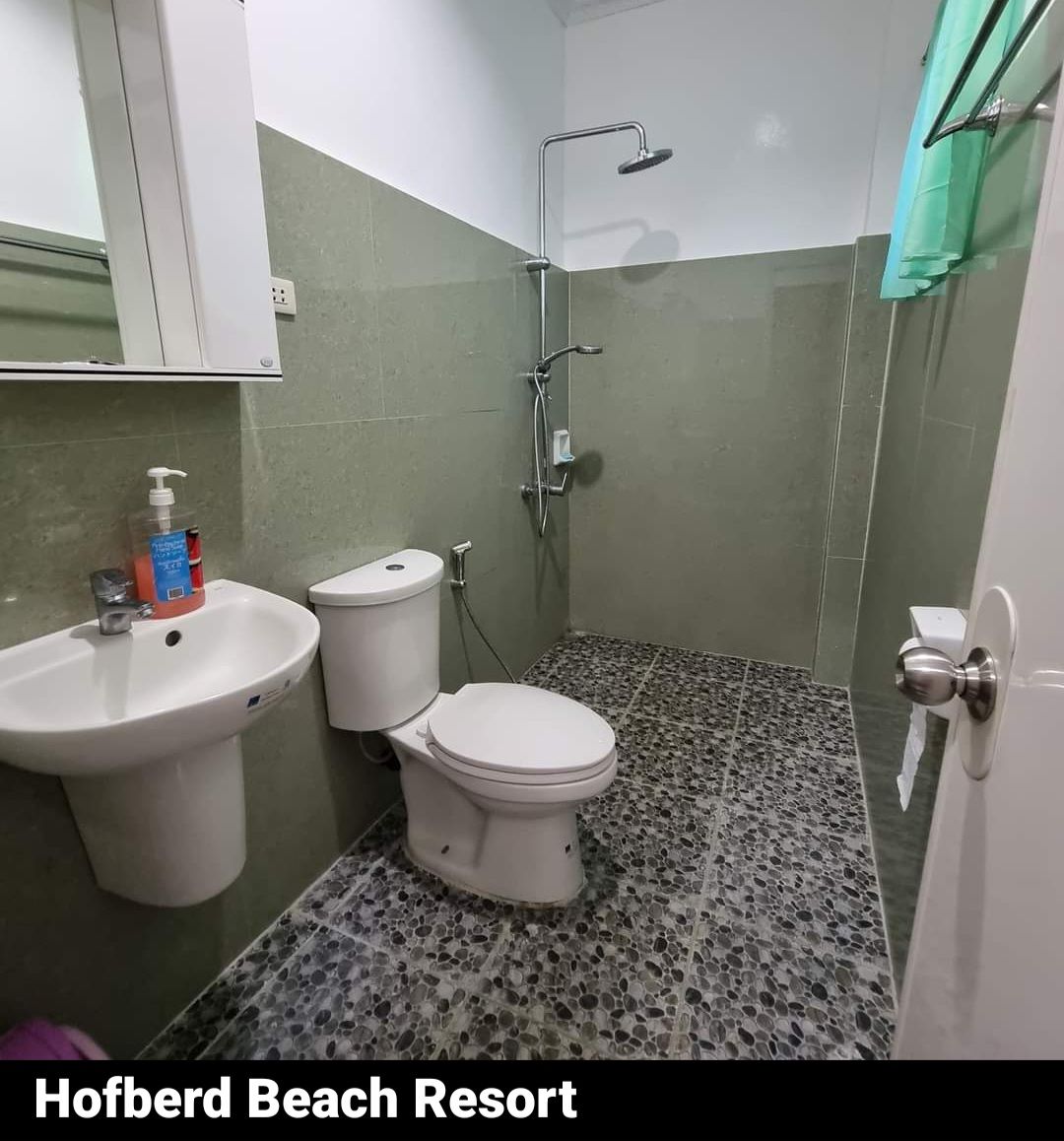 A functional bathroom is showcased with a shower area, a modern sink, and a toilet. The walls are tiled in muted green tones, complemented by a speckled stone floor. A small mirror hangs above the sink, while soft natural light is allowed in through a partially open window draped with green fabric.