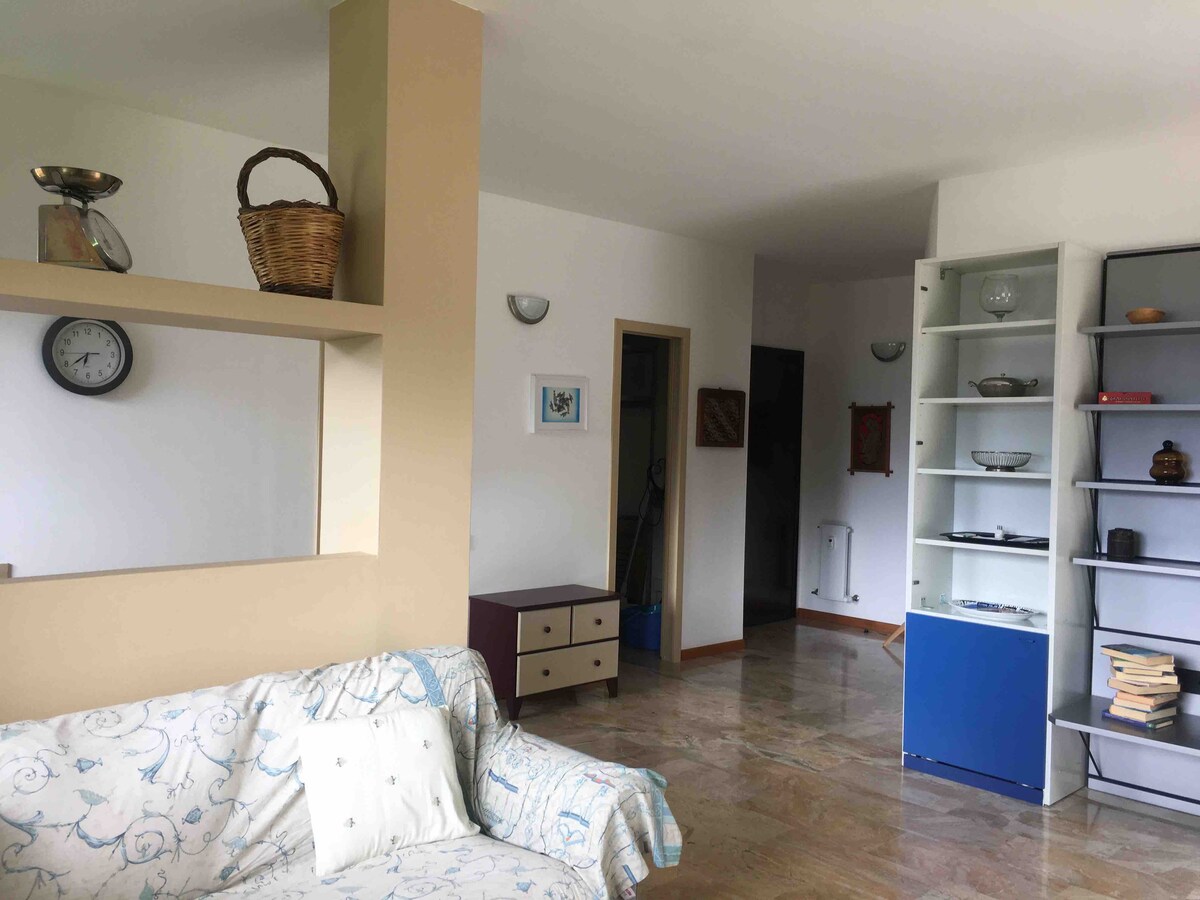 A cozy living area features a light-colored sofa with patterned details, complemented by a shelf unit displaying decorative items. A wall clock and a basket add functional charm, while a doorway leads to a hallway enhancing the room's open feel.