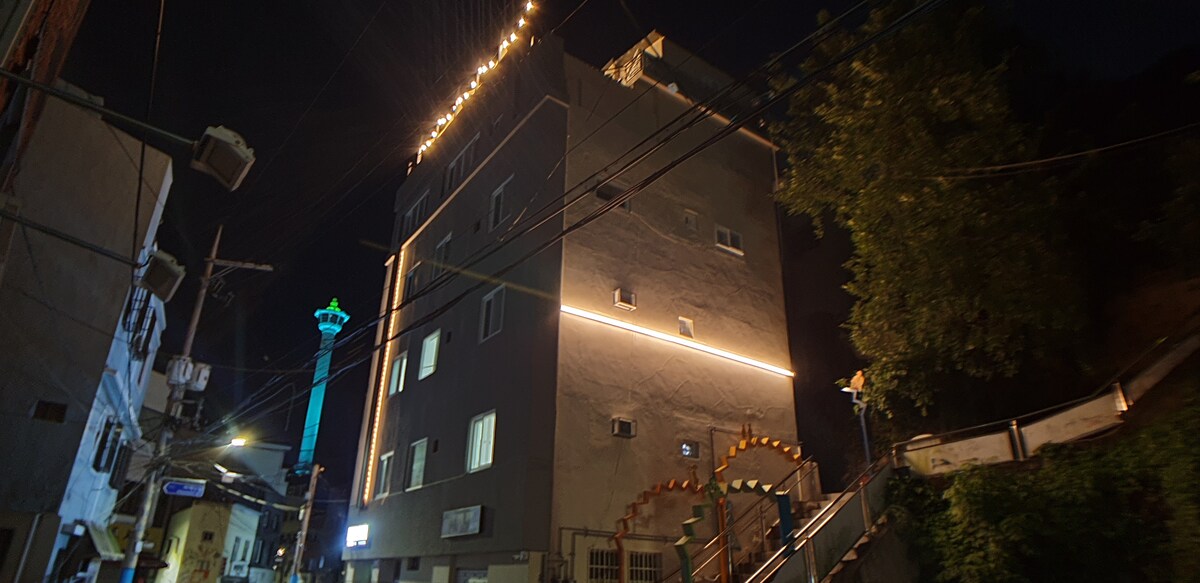 The exterior of a multi-story building is shown at night, highlighted by soft lighting along the walls. The upper part of the building features additional decorative elements. A tall green structure is visible in the background, adding a vibrant contrast to the scene.