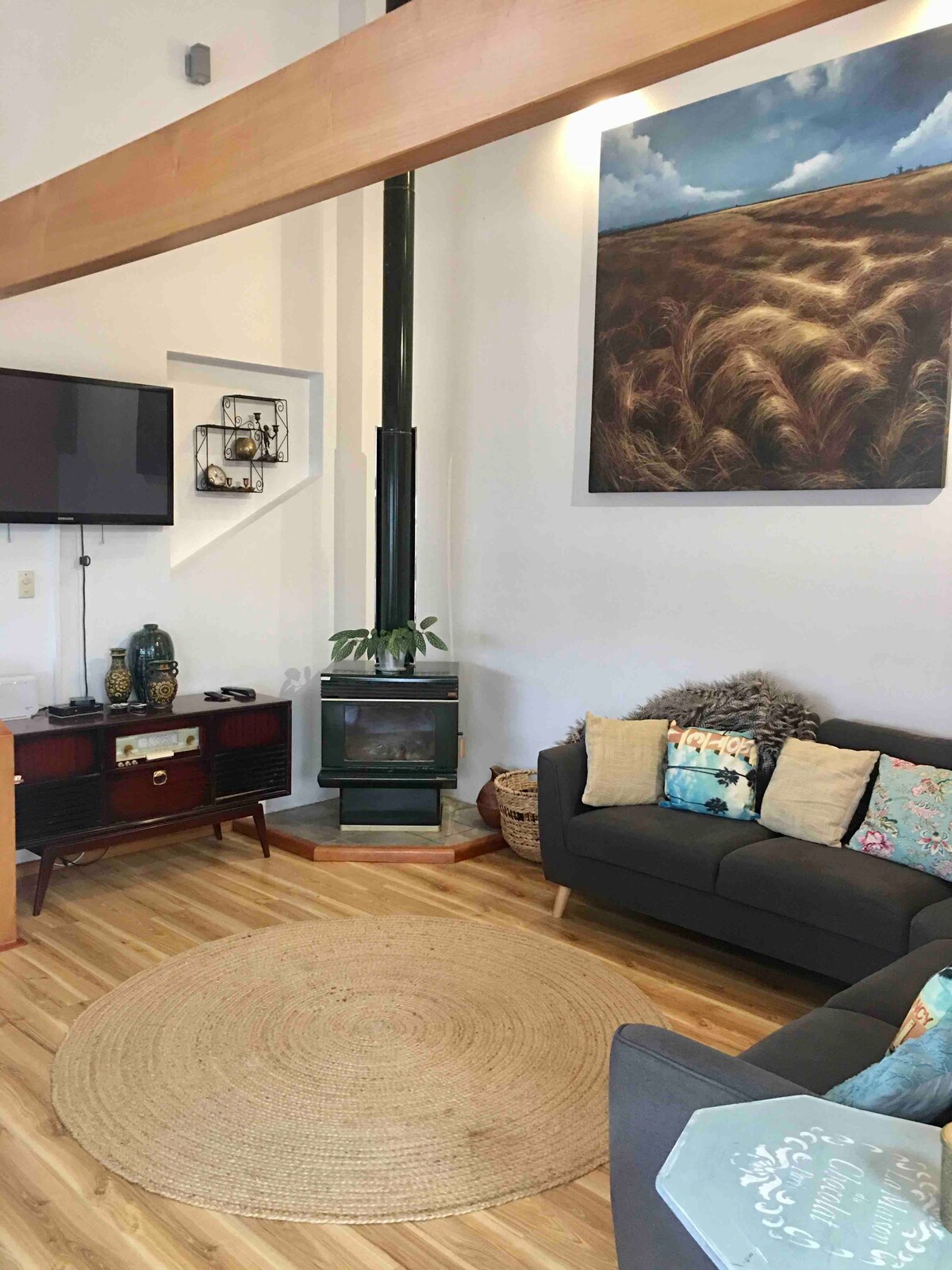 A modern living area is highlighted by a green wood stove and a large artwork depicting a serene landscape. Cozy seating includes a dark sofa adorned with patterned cushions. A natural fiber rug offers warmth to the wood floor, while a television is mounted on the wall.