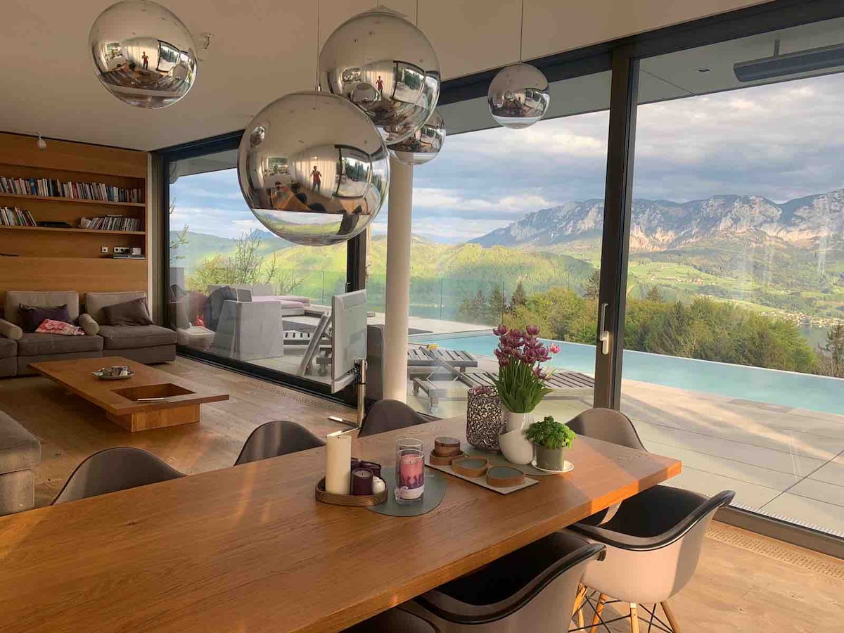 The spacious open-plan living and dining area features large glass doors that provide stunning views of the mountains and landscape. Modern pendant lights hang above a wooden dining table, complemented by stylish chairs. A cozy seating area is seen in the background, creating a harmonious space.