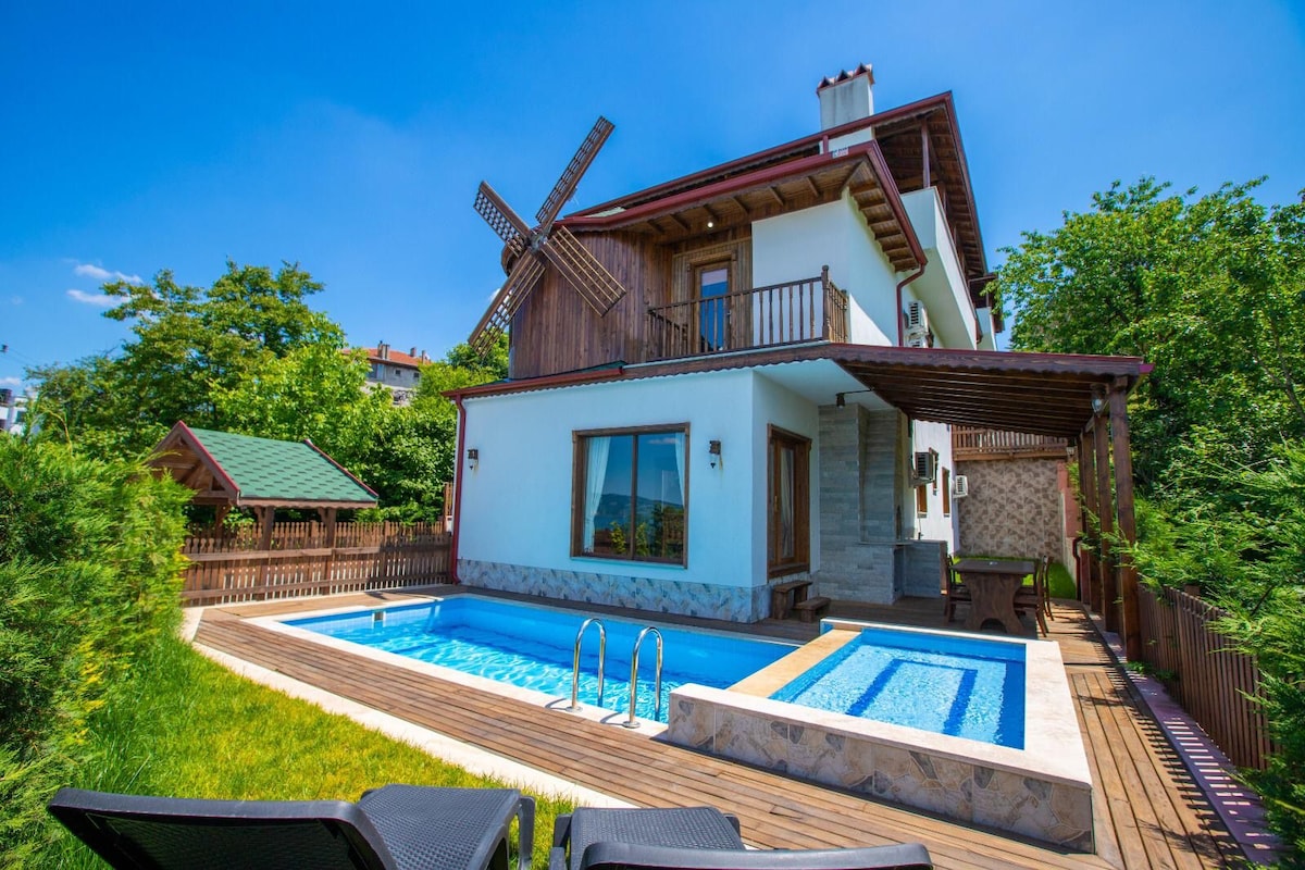The exterior of the villa is showcased, featuring a private pool surrounded by wooden decking. A traditional windmill adorns the roof. Lush greenery envelops the property, and a seating area is visible beneath a covered porch.